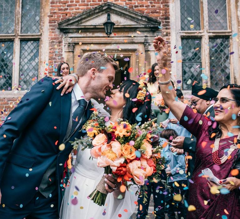 Bride in long sleeve mesh overlay wedding dress and colourful bridal flower crown kissing groom in classic navy grooms suit with black tie, grey waistcoat and colourful rose boutonniere amidst colourful wedding confetti exit 