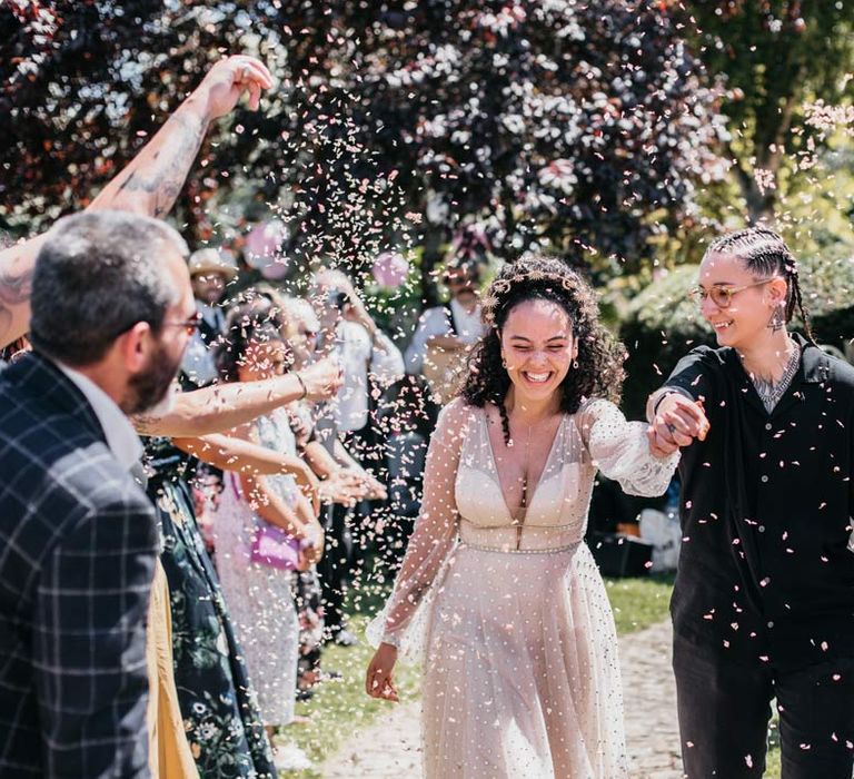 Bride in long sleeve v-neck sheer polka dot overlay sparkly wedding dress and gold celestial bridal headband holding hands with bride in black shirt and black trousers at Viking themed wedding doing colourful confetti exit