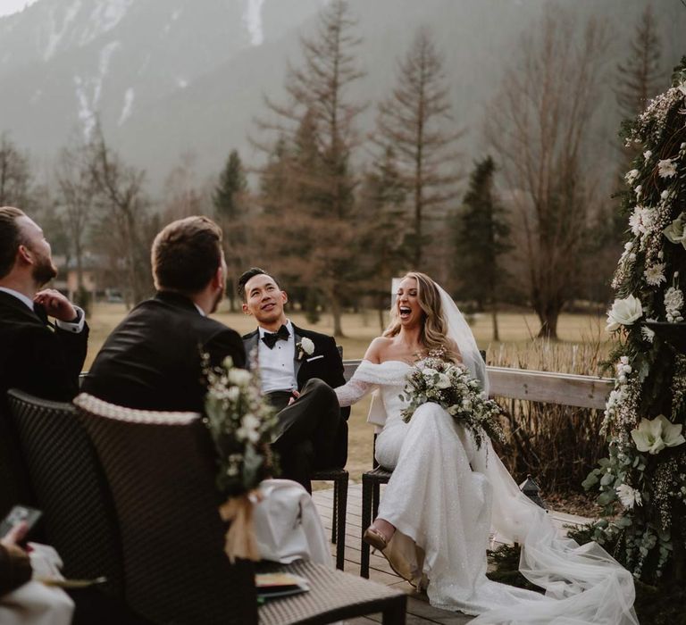 Groom in classic black tuxedo, black bowtie and white garden rose boutonniere and bride in off shoulder sparkly mermaid wedding dress holding white garden rose, eucalyptus, baby's-breath and foliage bridal bouquet laughing at the alter