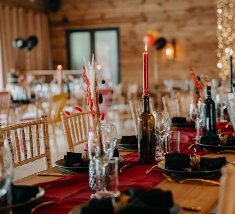 Gothic rock and roll wedding tablescape with deep red wedding table runner, black plates, black napkins, gold cutlery, red tapered candles in black wine bottles, and red white and black pampas grass wedding centrepieces at Southlands Barn