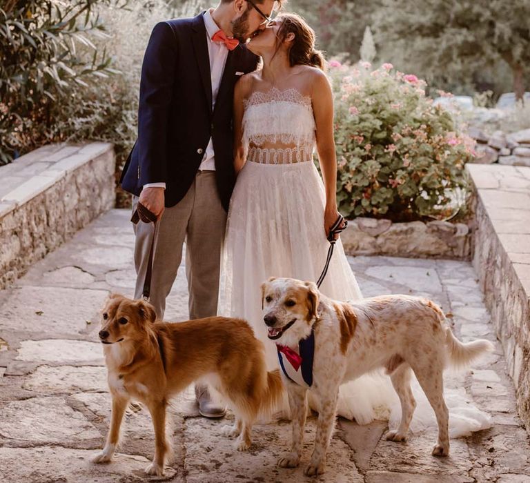 groom in grey chinos navy blazer and orange bow tie kissing his bride in a strapless lace wedding dress at greece destination wedding as they hold the leads of their dogs