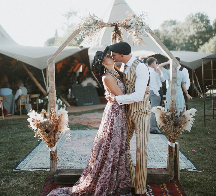 Bride wearing a pink boho wedding dress with silver wedding boots, aztec wedding jewellery and a boho bridal hat kisses groom wearing a mustard pinstripe suit, lilac socks, black sunglasses and a black hat in front of tipi wedding entrance surrounded by moroccan rugs and dried flower decor