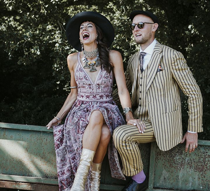 Bride wearing a pink boho wedding dress with silver wedding boots, aztec wedding jewellery and a boho bridal hat sits next to groom wearing a mustard pinstripe suit, lilac socks, black sunglasses and a black hat