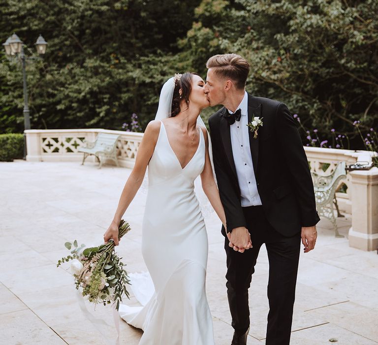 Bride in plunging satin slip wedding dress with cathedral length veil kissing groom in black tie 