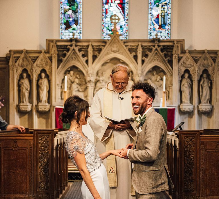 The bride in a beaded wedding dress and groom in a grey suit smile and laugh during their ceremony 