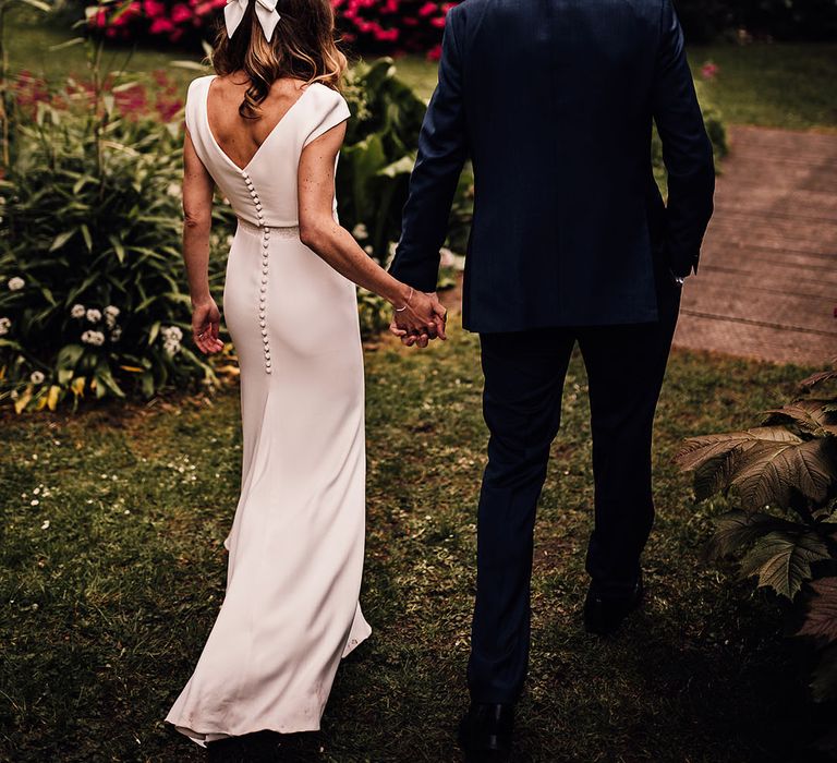 Bride in a button back wedding dress with white hair bow walking hand in hand with the groom in a blue suit 