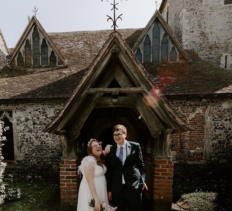 Bride wearing feather wedding dress with funky sunglasses and headband posing with the groom with purple accessories 