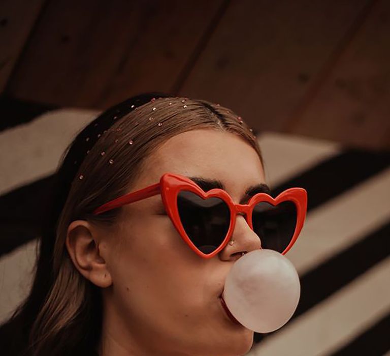 Woman wearing red love heart sunglasses blowing a bubble gum bubble by Marilyn Denise Photography