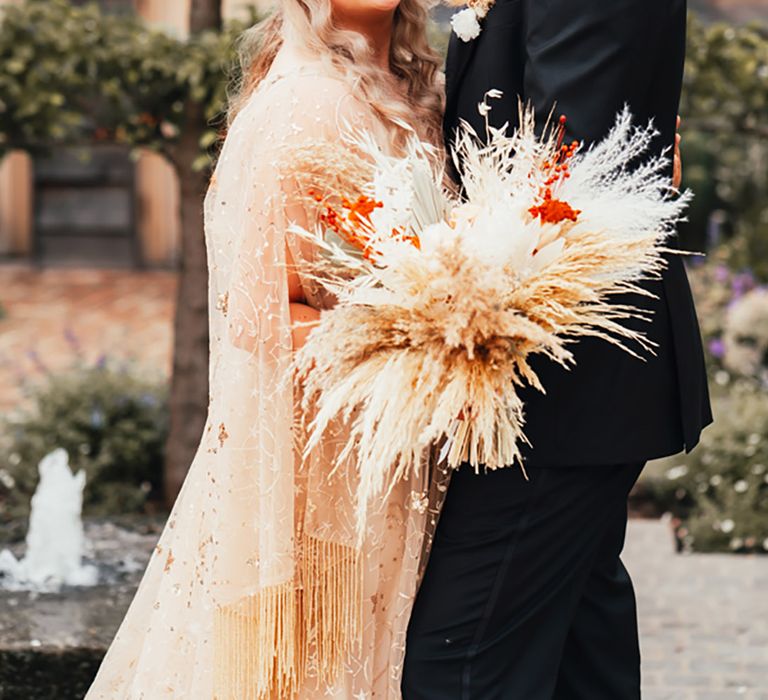 Bride in gold sparkly wedding dress with pampas grass wedding bouquet with the groom in an all black wedding outfit 