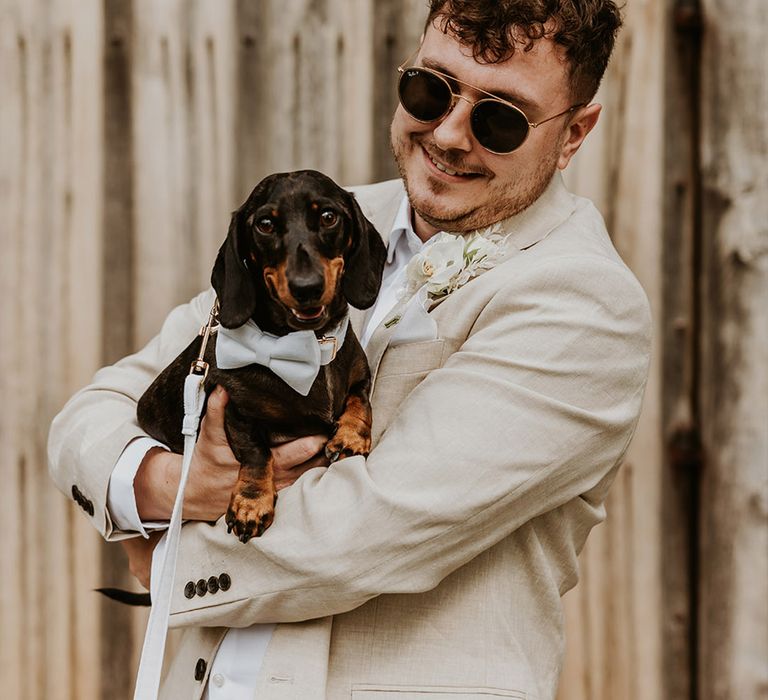 Groom in neutral beige wedding suit holding pet dog wearing a silver bow wedding collar 