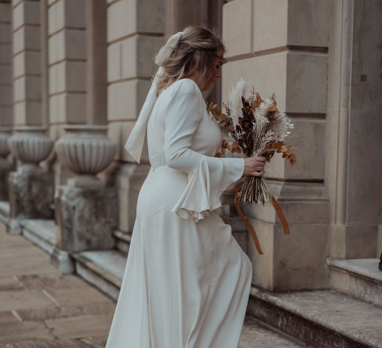 Bride walks up the stairs at Cliveden House in Ghost dress and holding dried floral bouquet 