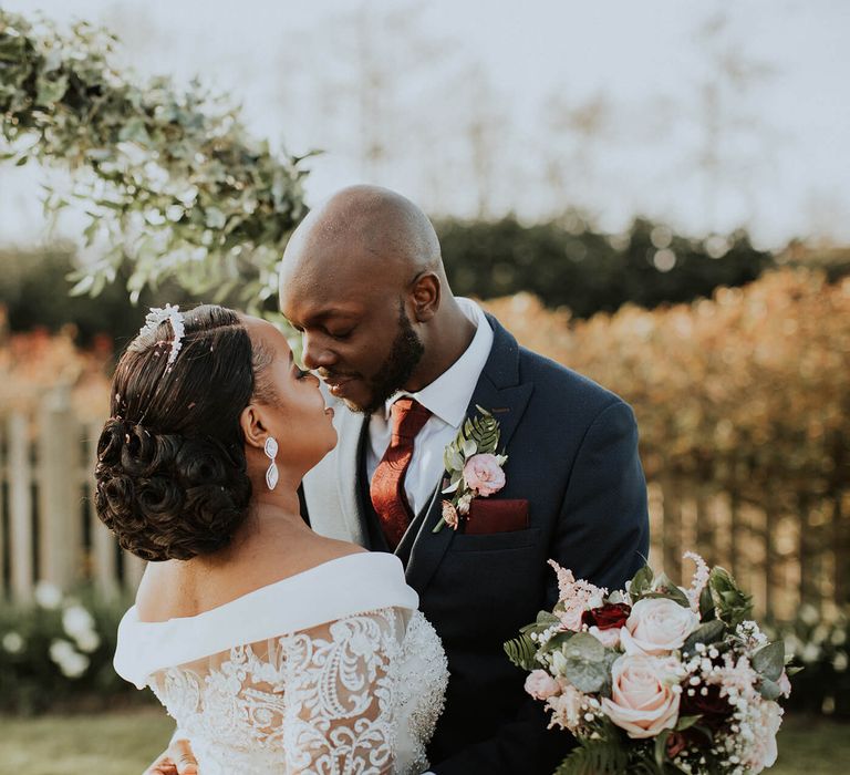 A Black couple embrace for wedding photos. The bride wears an off the shoulder dress and has her hair in a low bun.