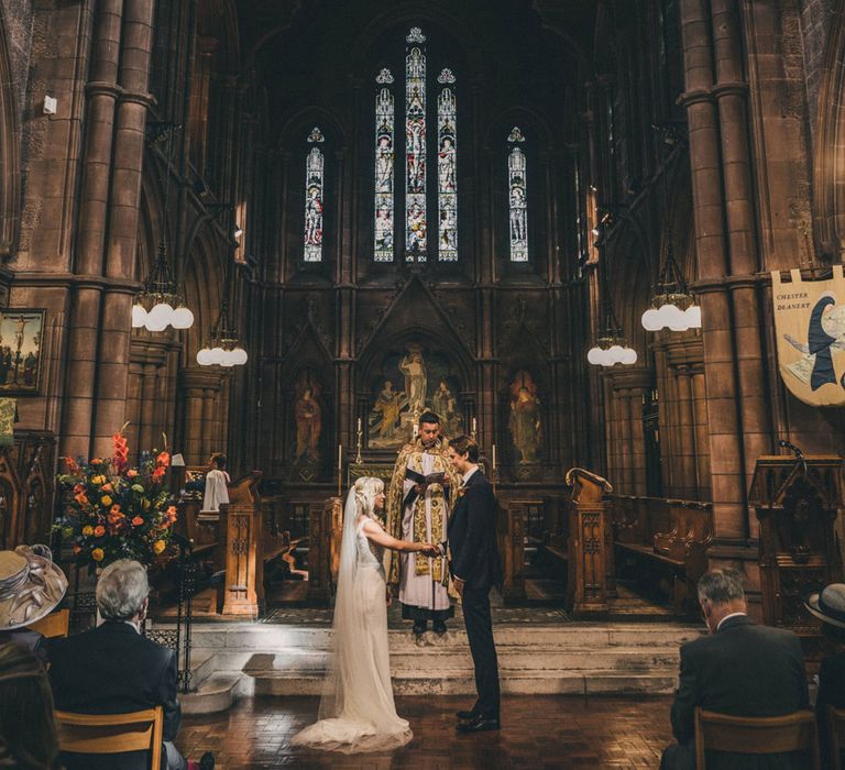 Bride in floor length veil and Justin Alexander wedding dress holds hands with groom at the altar whilst priest conducts ceremony