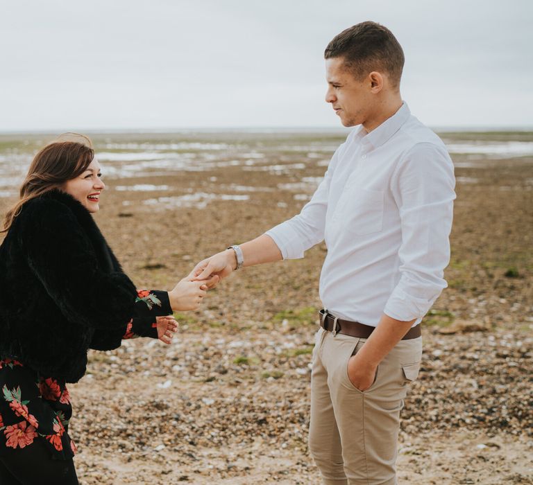 Groom-to-be in a white shirt and beige chino's holding his bride-to-be's hand on the beach during an engagement photo shoot