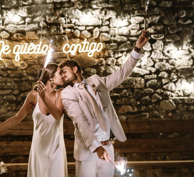 Bride in a slip wedding dress and groom in a beige suit holding sparklers in front of a stone wall with a neon sign 