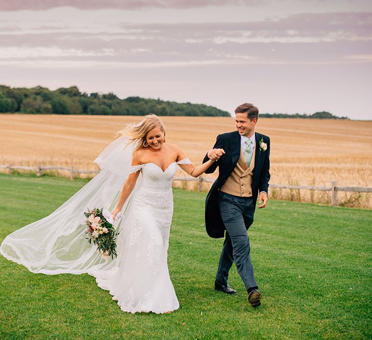 Bride and groom walking through countryside, bride wearing cold shoulder wedding dress and groom wearing three-piece suit