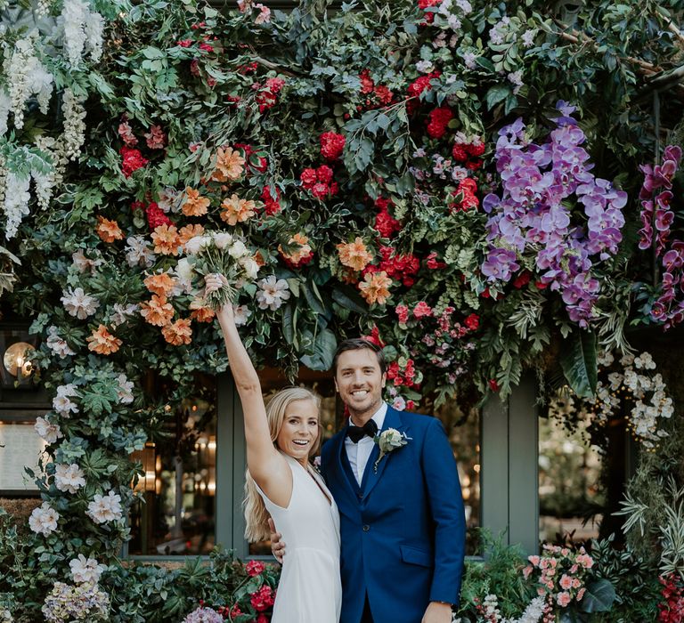 Bride & groom stand in front of colourful flower wall after wedding ceremony 