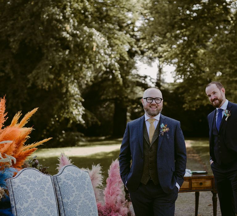 Groom in blue suit with green waistcoat and yellow tie stands waiting for bride with hands in pockets at pink pampas grass wedding