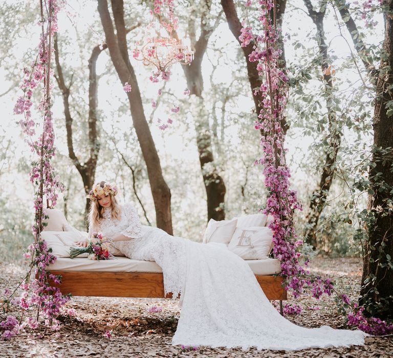 Bride in long sleeved lace boho wedding dress and flower crown lounges on large wooden swinging day bed with pink floral decor at enchanted forest wedding