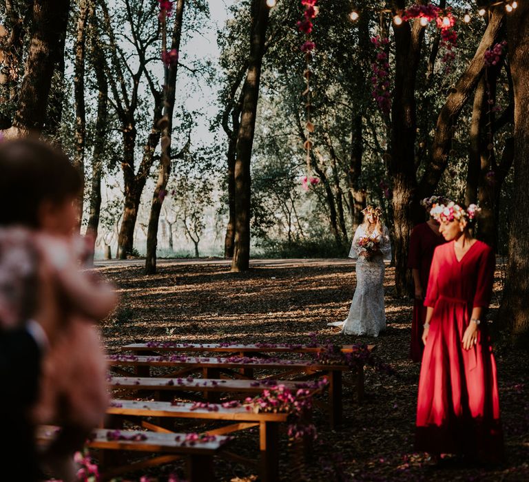 Bride in long sleeved lace boho wedding dress walks down woodland wedding aisle behind bridesmaid in long fuchsia bridesmaid dress and flower crown at enchanted forest wedding in Italy 