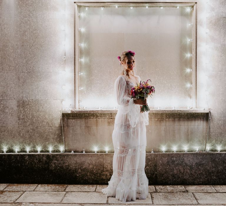 Smiling bride with curled hair, pink floral hair accessories and long tulle sleeved boho wedding dress holds multicoloured wedding bouquet whilst standing in front of fairy lights at enchanted forest wedding in Italy