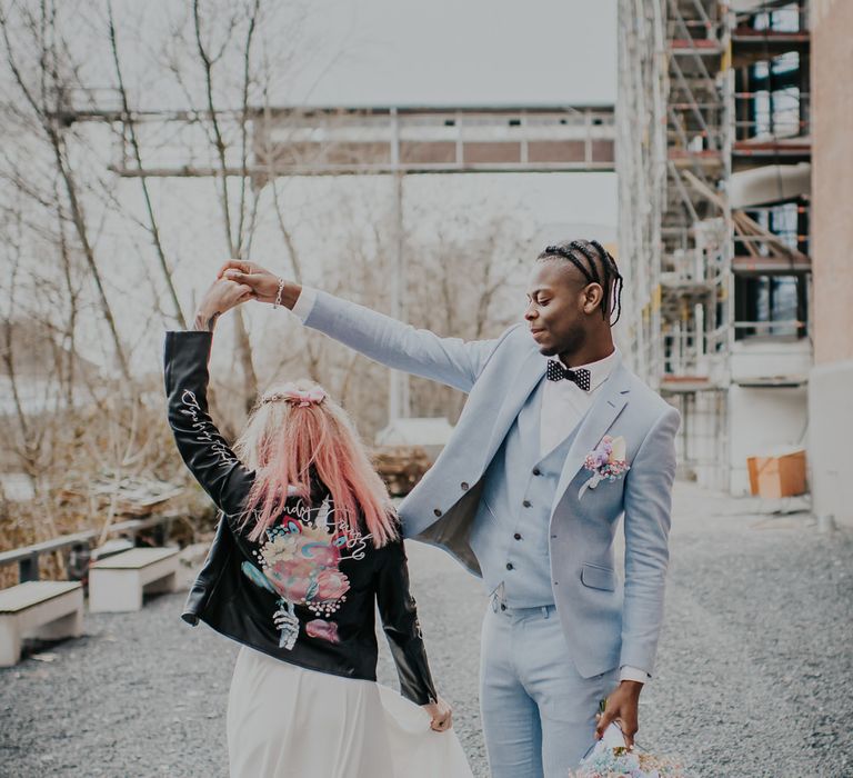 Groom in a pale blue wedding suit holding a pastel bouquet twirling his bride in a customised leather jacket 