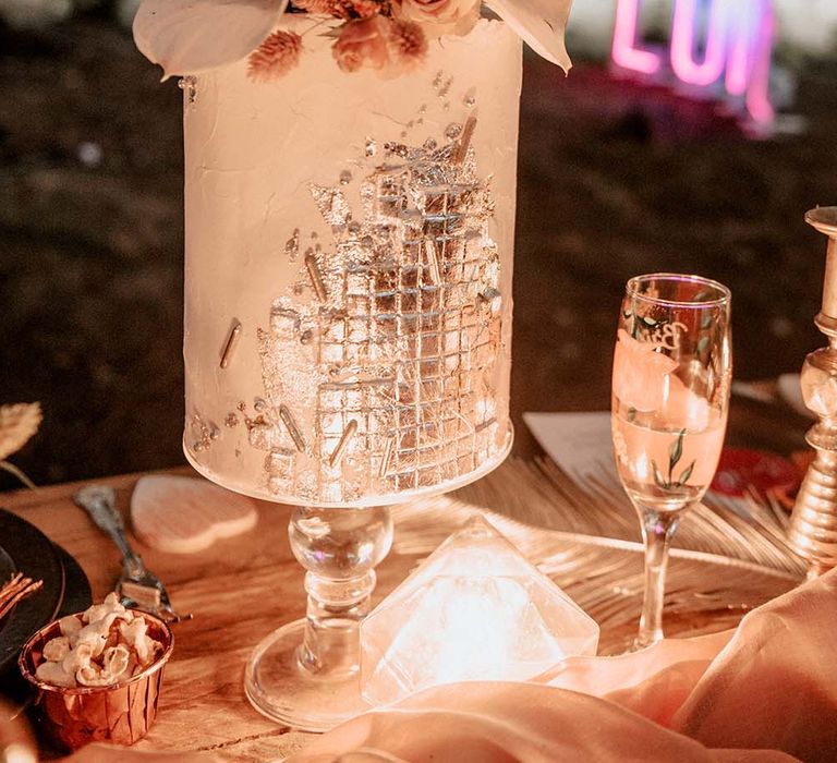 White column wedding cake with silver disco ball detailing and topped with tropical flower decor on glass cake stand at festival themed wedding