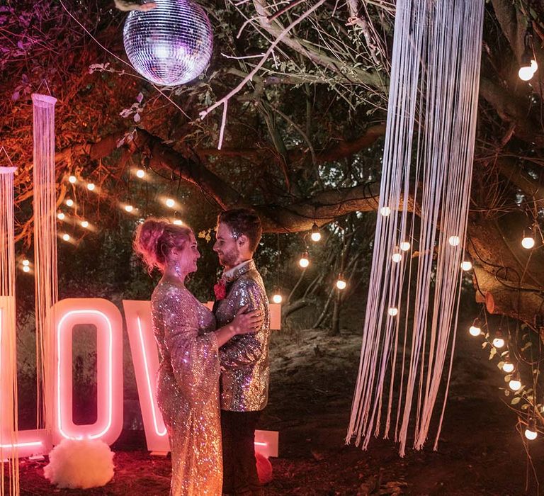 Bride with pink curled hair and gold sequinned dress dances with silver sequin suit jacket under disco ball and by pink neon LOVE sign outside at festival themed wedding