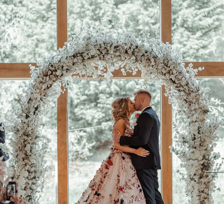 Bride & groom kiss beneath floral archway on their wedding day