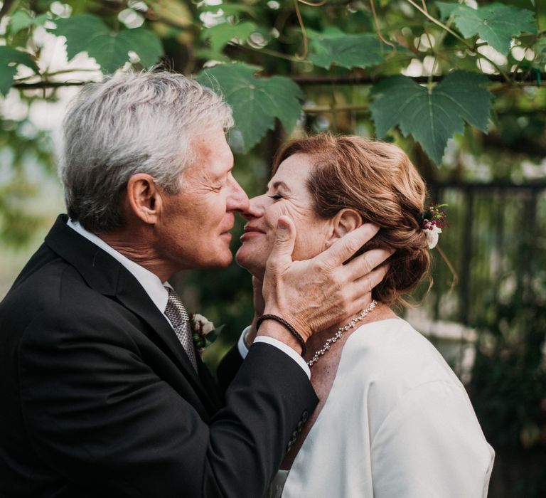 Groom holds the face of his wife as he leans in for a kiss whilst outdoors in Italy surrounded by green foliage