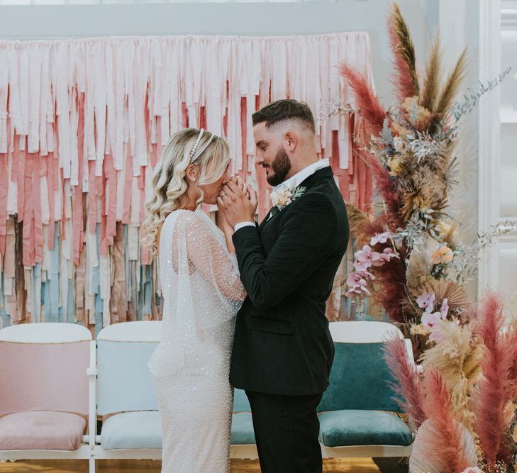 Bride in a sparkly wedding dress holding hands with her groom in front of a pastel tissue tassel wall back drop with vertical flower arrangements 