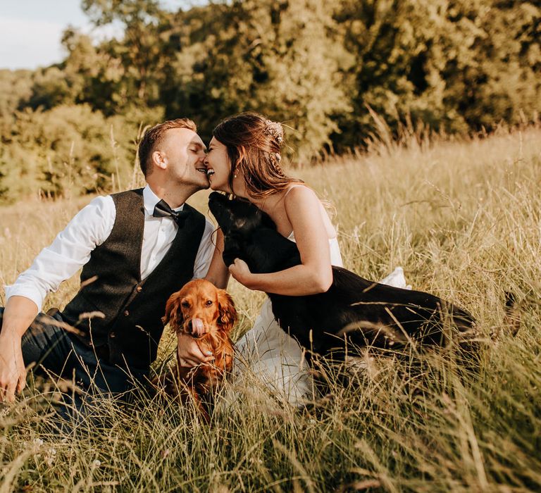 Bride in white cami dress with flowers in her hair and groom in waistcoat and white shirt with rolled up sleeves sit together and kiss in field with two dogs at garden wedding reception