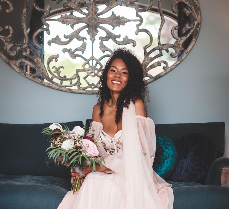 Black bride with naturally curly hair in blush bridal separates with an applique veil sitting on a sofa holding a deep red. pink, white and green bouquet