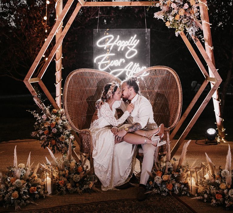 Bride and groom sitting on peacock chairs under a hexagonal wooden frame surrounded by dried and fresh flower arrangements 