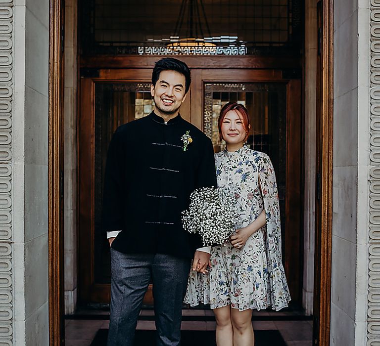 East Asian bride and groom standing outside Old Marylebone Town Hall in a navy jacket and blue floral dress holding a gypsophila wedding bouquet 