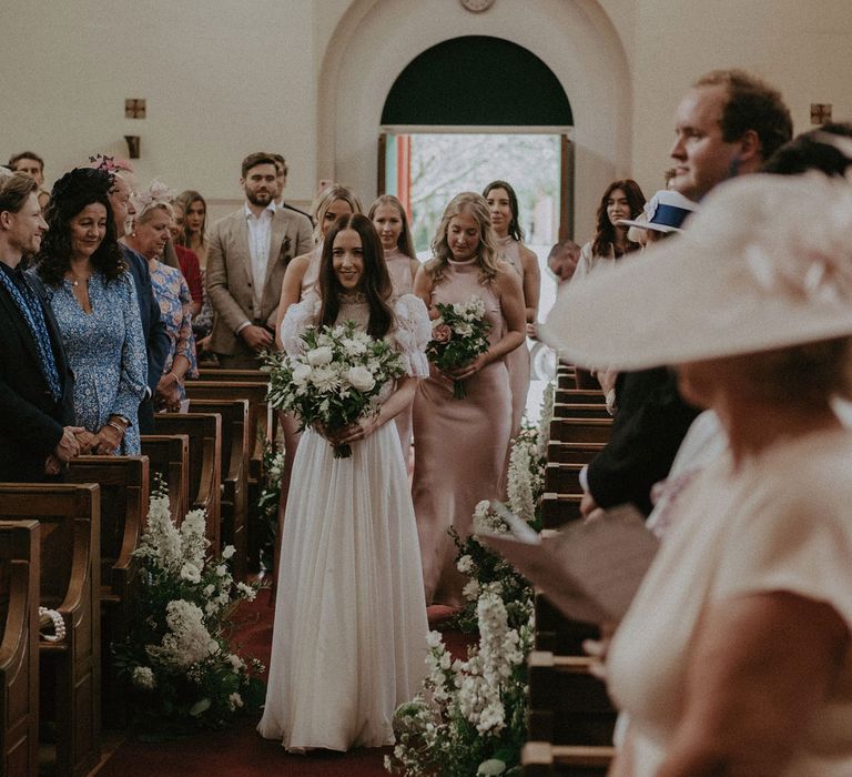 Bride in white lace puffed sleeved Daalarna dress holding white and green bridal bouquet walks down the aisle during church ceremony whilst bridesmaids in blush pink satin bridesmaids dresses follow behind 