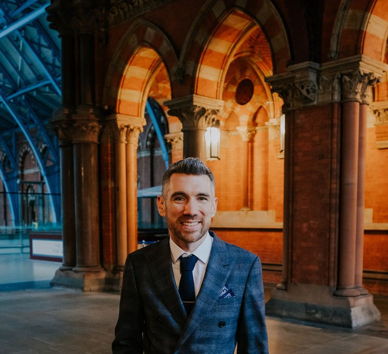 Groom stands within St Pancreas as he holds his hand within his trouser pocket and wears grey suit complete with burgundy tie