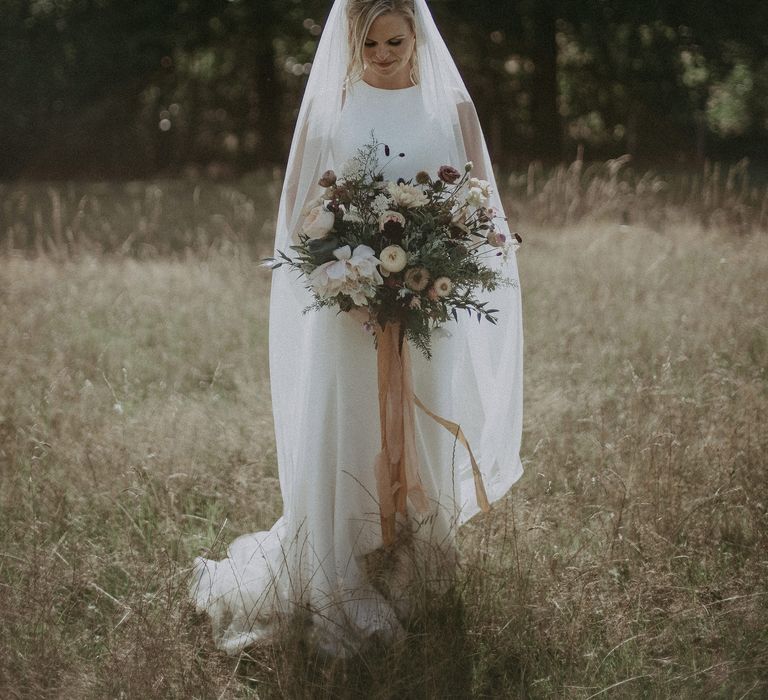 Bride stands front facing as she holds her floral bouquet and looks down toward the ground whilst smiling