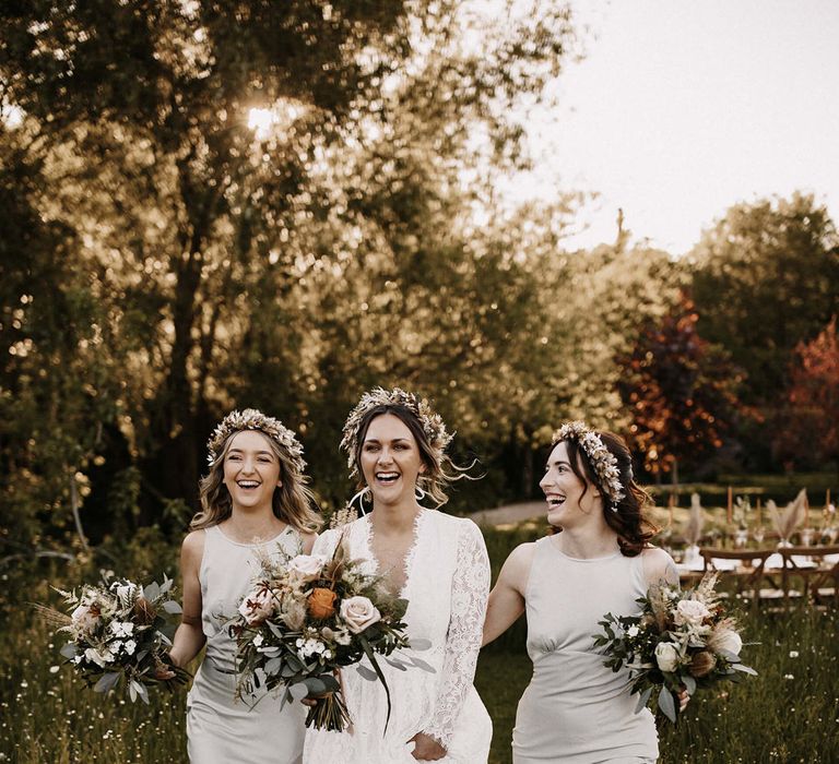 Bride in a lace long sleeve wedding dress with her two bridesmaids in silver satin dresses and all wearing dried flower crowns 