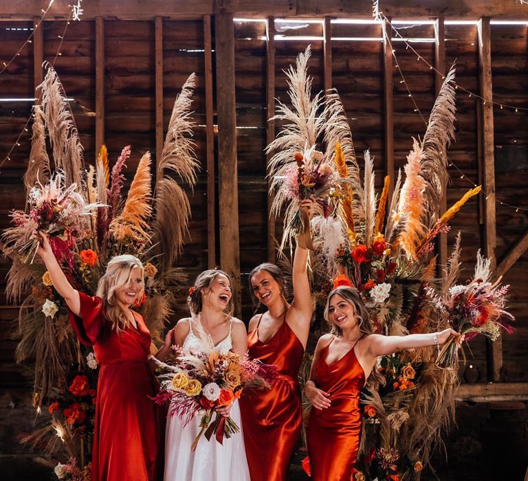 Bride and bridesmaids in deep red satin bridesmaid dresses standing in a rustic barn with dried flower arrangements 