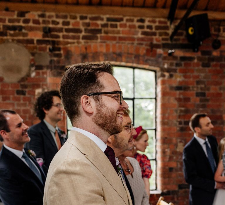 Groom in double breasted linen suit smiles whilst waiting for bride to walk up the aisle during wedding ceremony at Loft Studios London