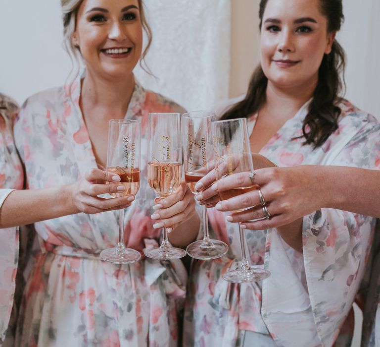 Bride and bridesmaids in floral bridal gowns clinking personalised flutes before Tythe Barn wedding with barn wedding flowers