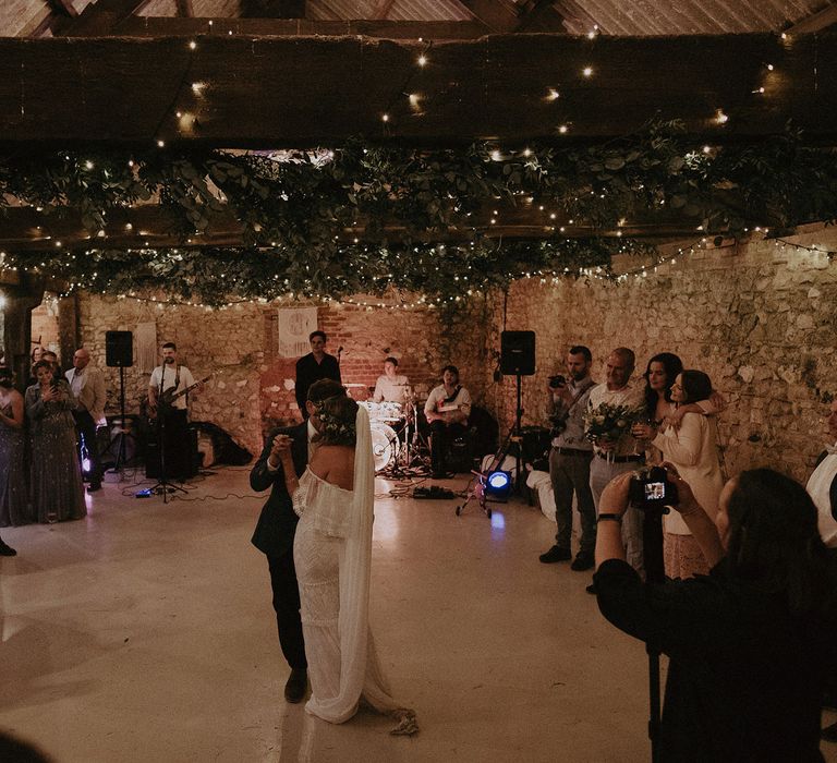 Bride in Grace Loves Lace bardot wedding dress and veil dances with groom in blue suit jacket under fairy light and foliage canopy as guests watch on at Isle of Wight wedding with macrame wedding decor