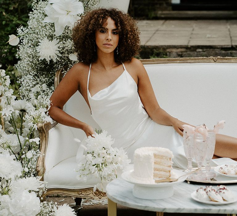 Black bride with naturally curly hair in a satin slip wedding dress lounging on a sofa 