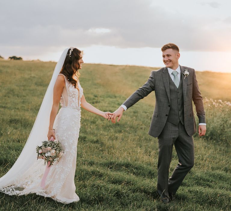 Bride in applique Berta wedding dress, headband and veil holding mixed bridal bouquet walks through field holding hands with groom in grey there piece checked suit during summer wedding at Primrose Hill Farm