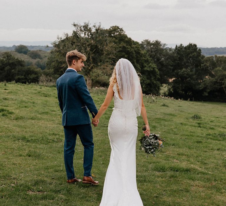 Bride & groom walk across green fields in Sussex on their wedding day