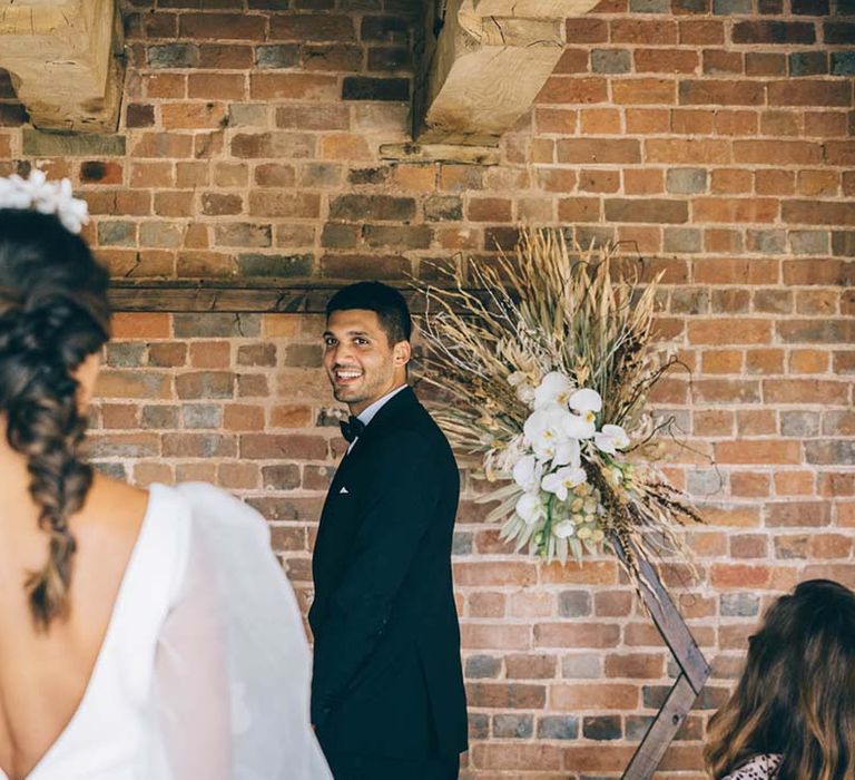 Groom in a tuxedo standing at the altar smiling at his bride as she walks down the aisle at their boho black tie wedding 