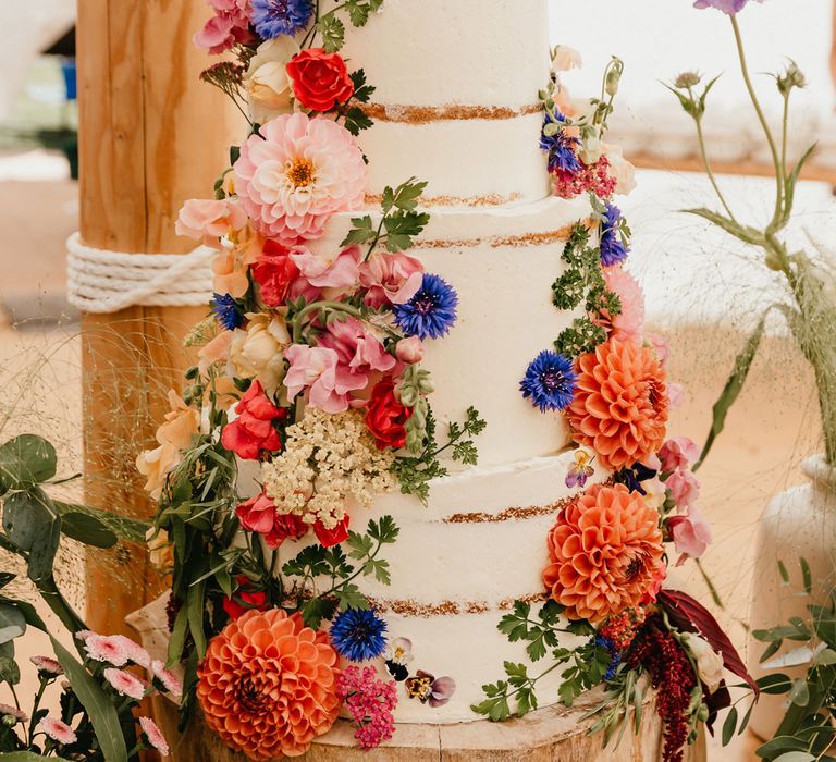 Semi naked wedding cake on a rustic tree slice cake stand decorated with colourful dahlia wedding flowers 