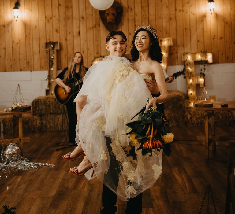 Bride in feathered tulle wedding dress and red heels holding bouquet is held by groom in dark suit during rustic wedding ceremony with fairy lights