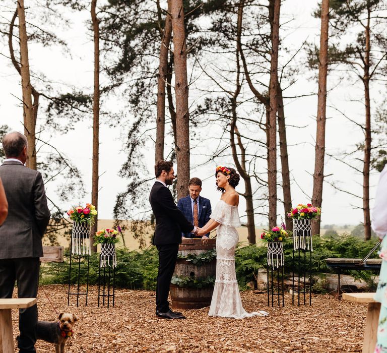 Bride in Grace Loves Lace wedding dress and flower crown stands holding hands with groom in black suit during woodland wedding ceremony at The Bridal Barn wedding venue